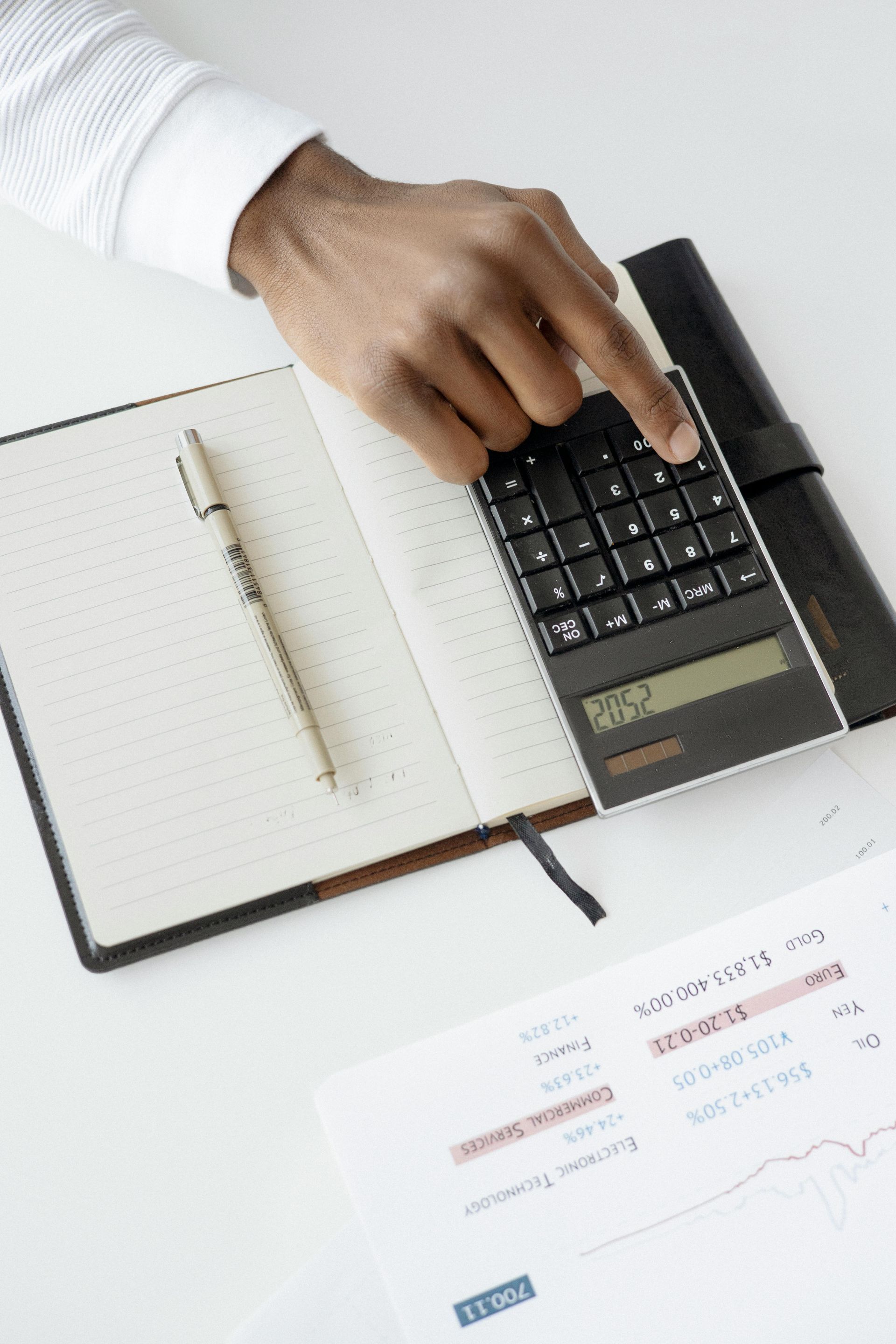 A person's hand pressing a calculator button next to an open notebook, a pen, and financial documents on a white desk.