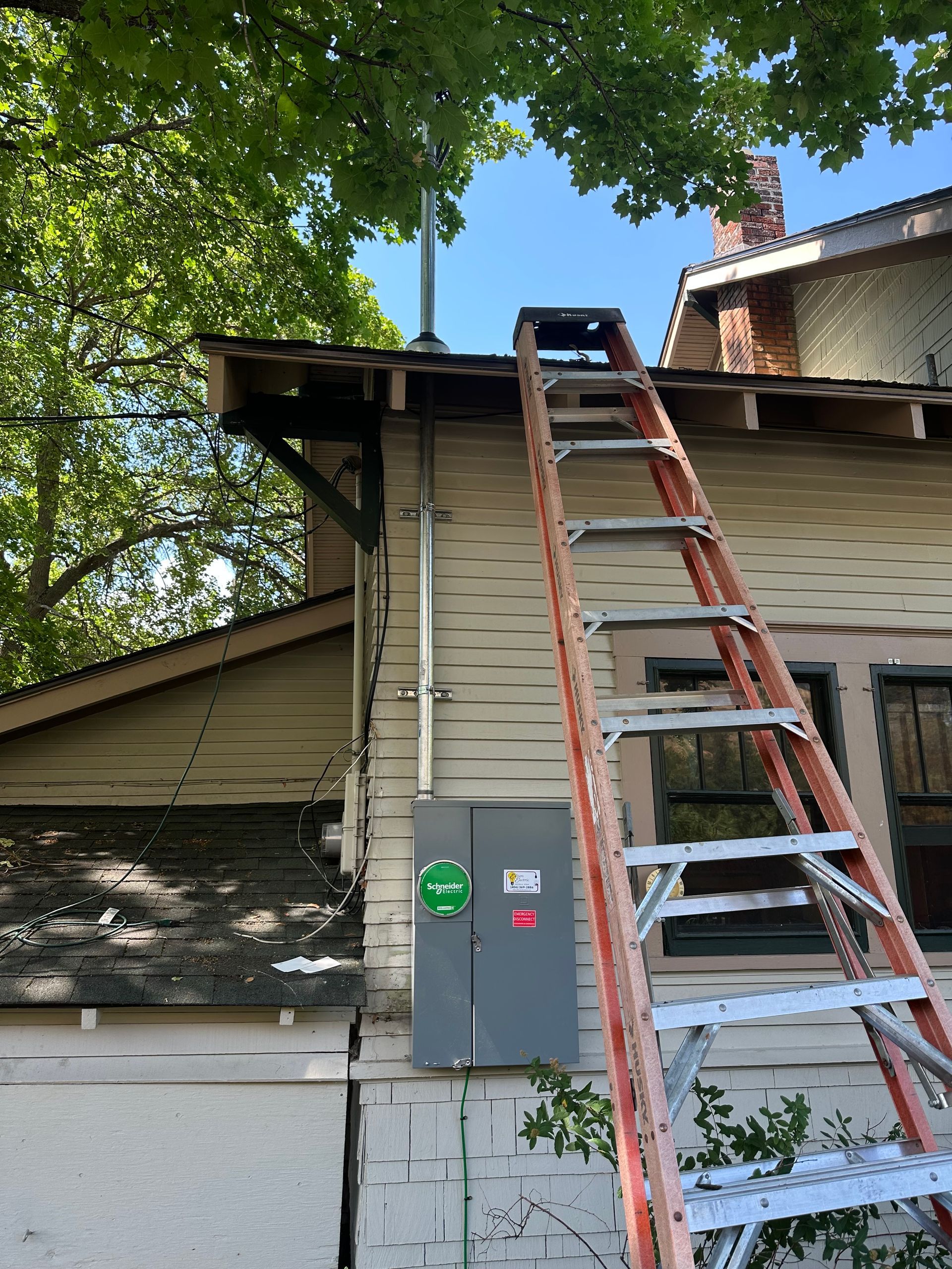 Ladder leaning against a house roofline beside a gray utility box and leafy trees