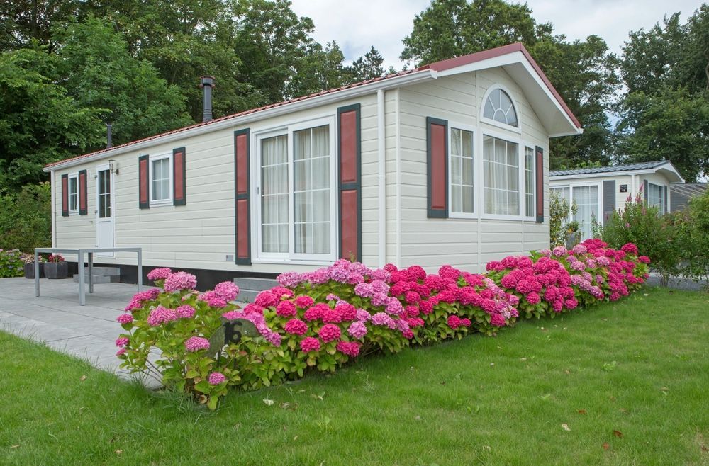 Small white mobile home with red shutters and pink flowers along the front lawn