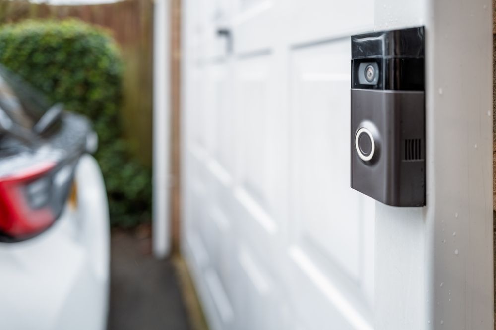 Black video doorbell mounted on a white front door, with a blurred car in the driveway.