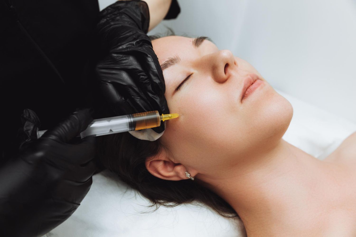 Woman receiving facial injection; gloved hand holds syringe near cheek in medical setting.