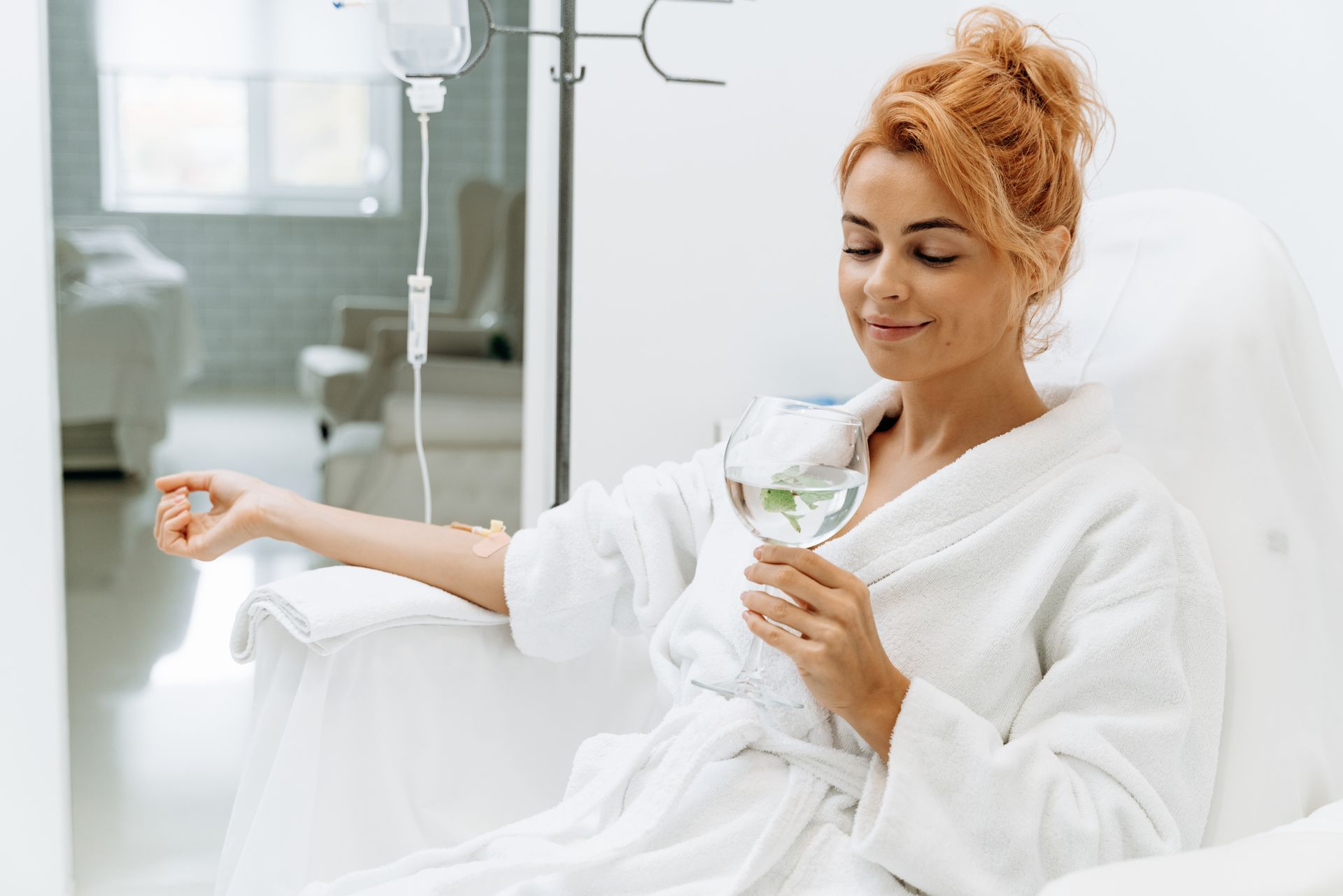 Woman in white robe, receiving IV drip, holding glass of water, sitting in a chair in a spa-like setting.