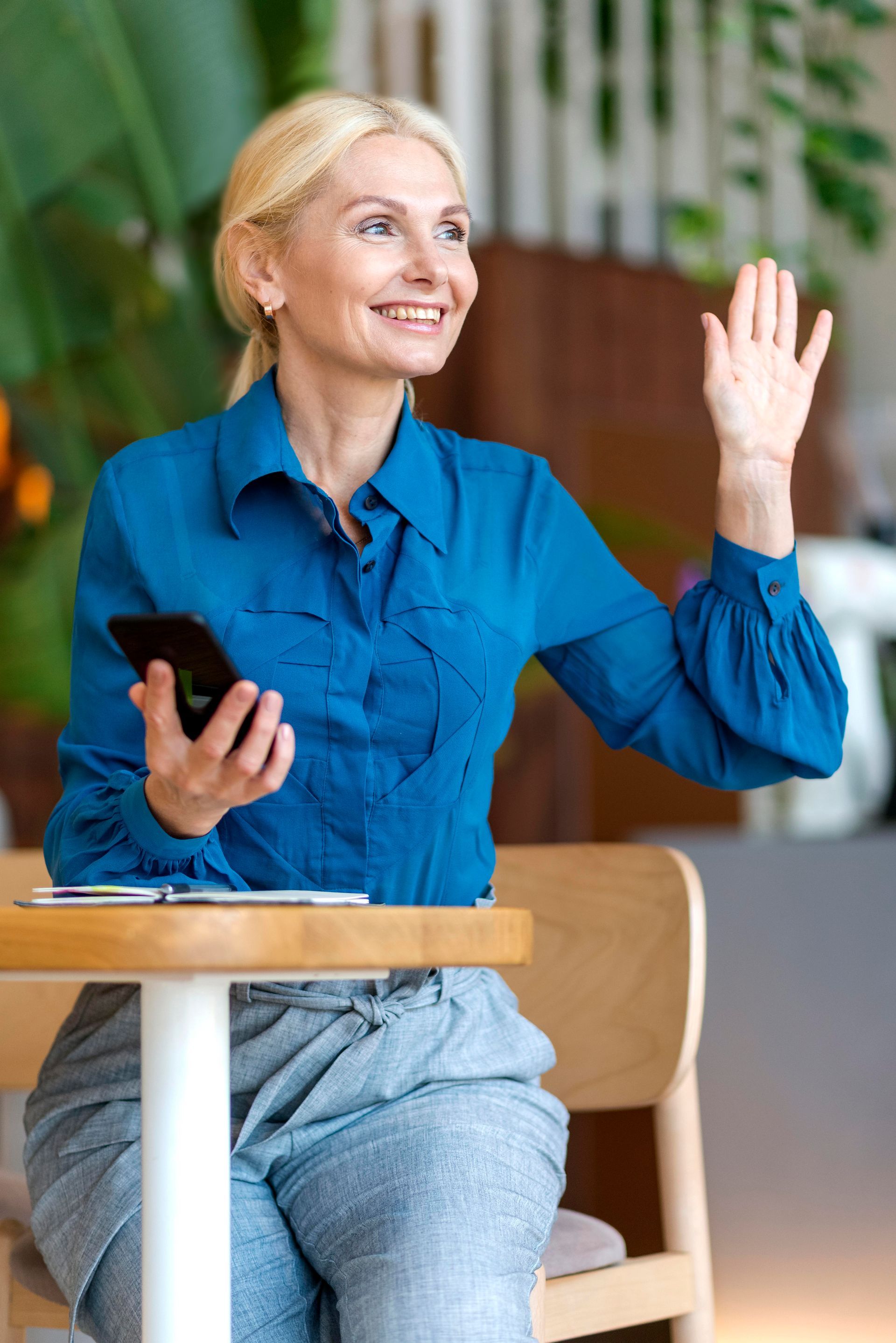 a woman is sitting at a table holding a cell phone and waving .