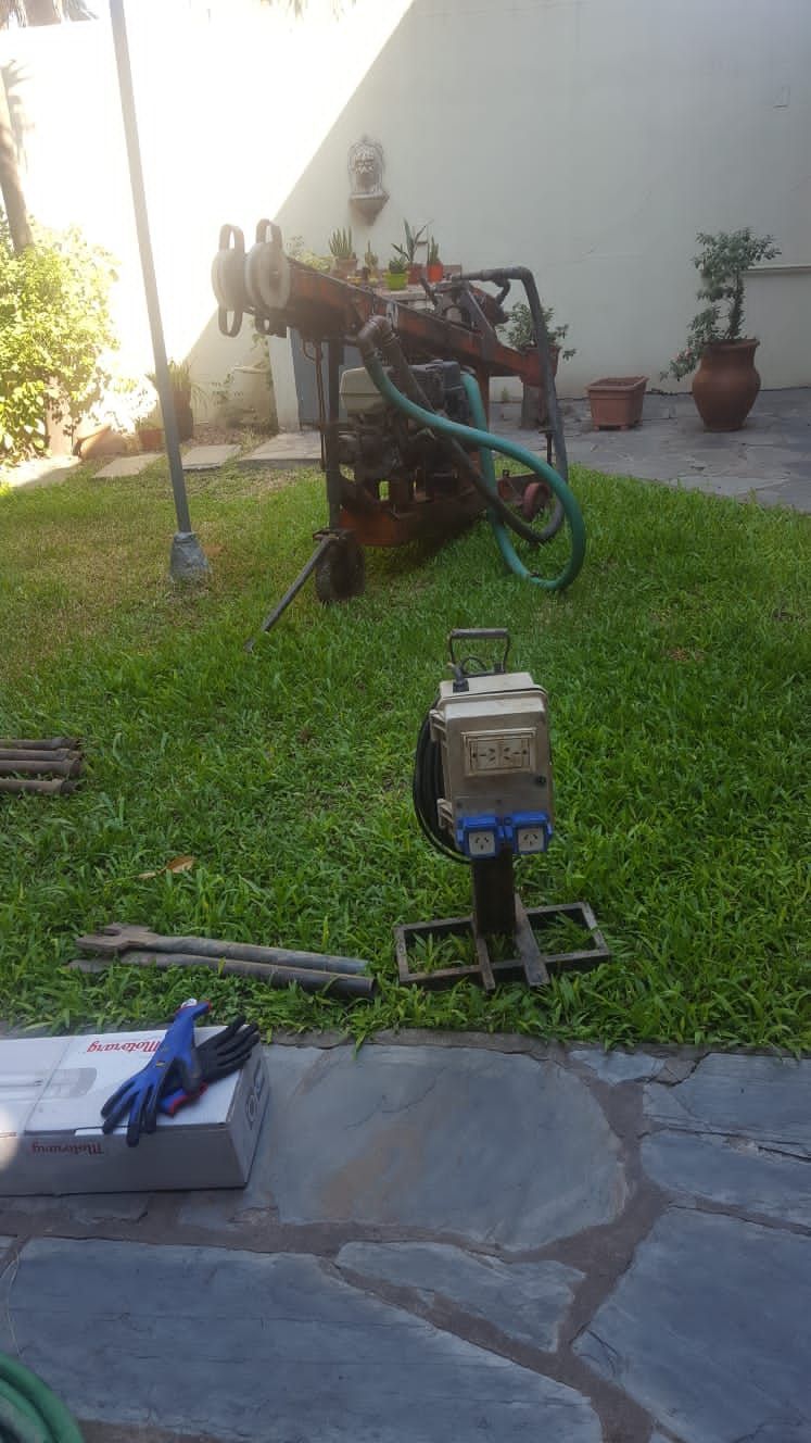 Pequeño jardín con una máquina azul sobre un soporte, un patio de piedra, una manguera y plantas en macetas cerca de una pared.