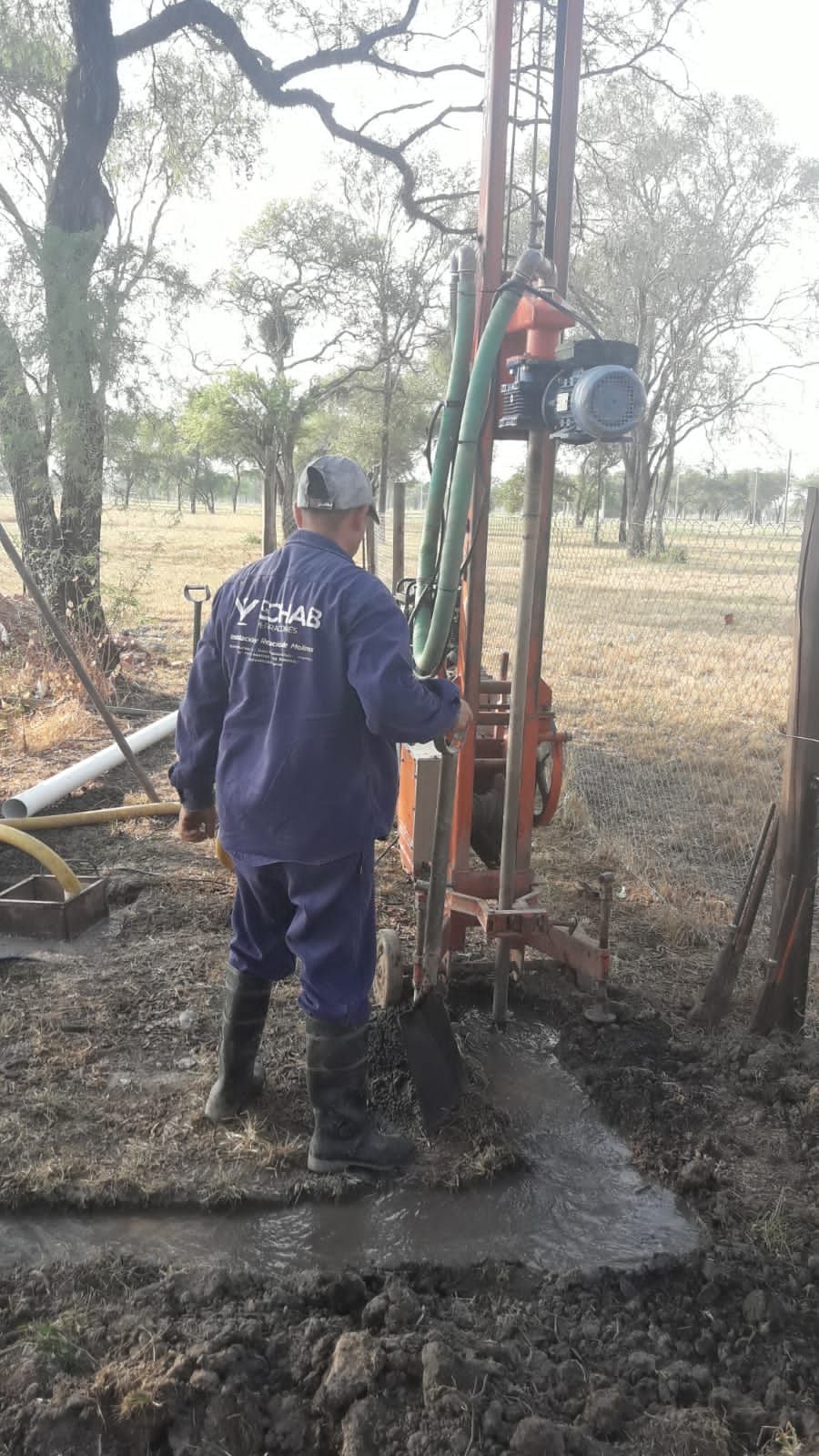 Un trabajador perfora un pozo de agua en un campo fangoso con una plataforma de perforación y árboles cerca.