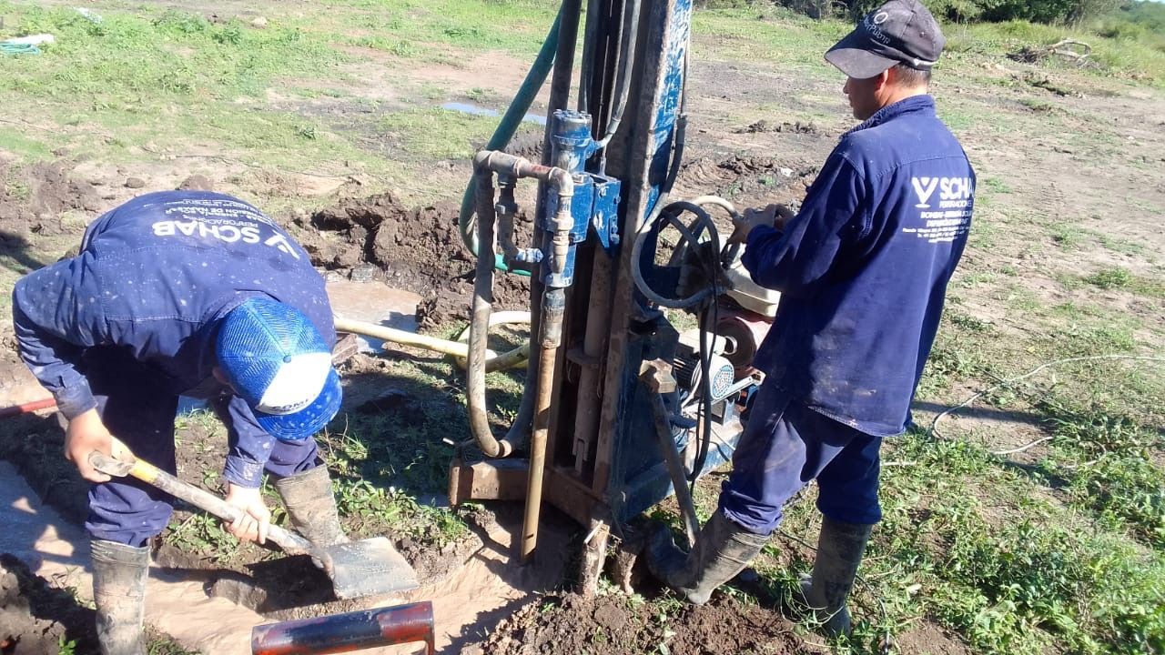 Dos trabajadores con uniformes azules excavan alrededor de una perforadora en un campo de hierba.