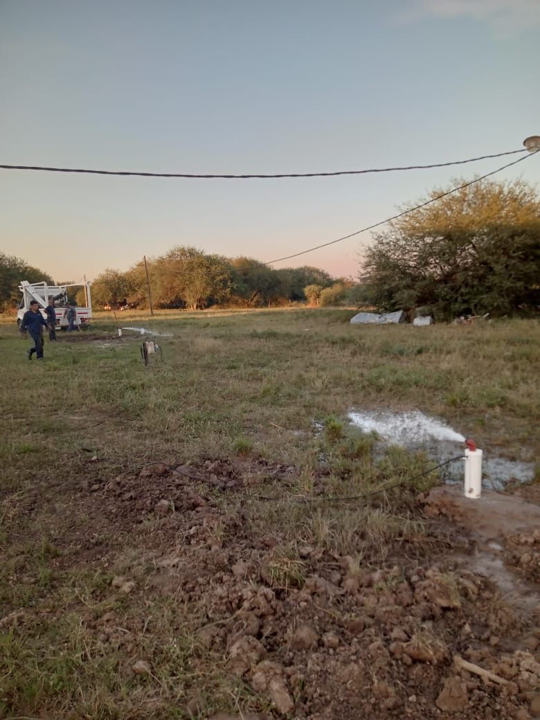 Campo rural al atardecer con una persona cerca de un perro blanco, líneas eléctricas aéreas y una pequeña tubería de agua en primer plano.