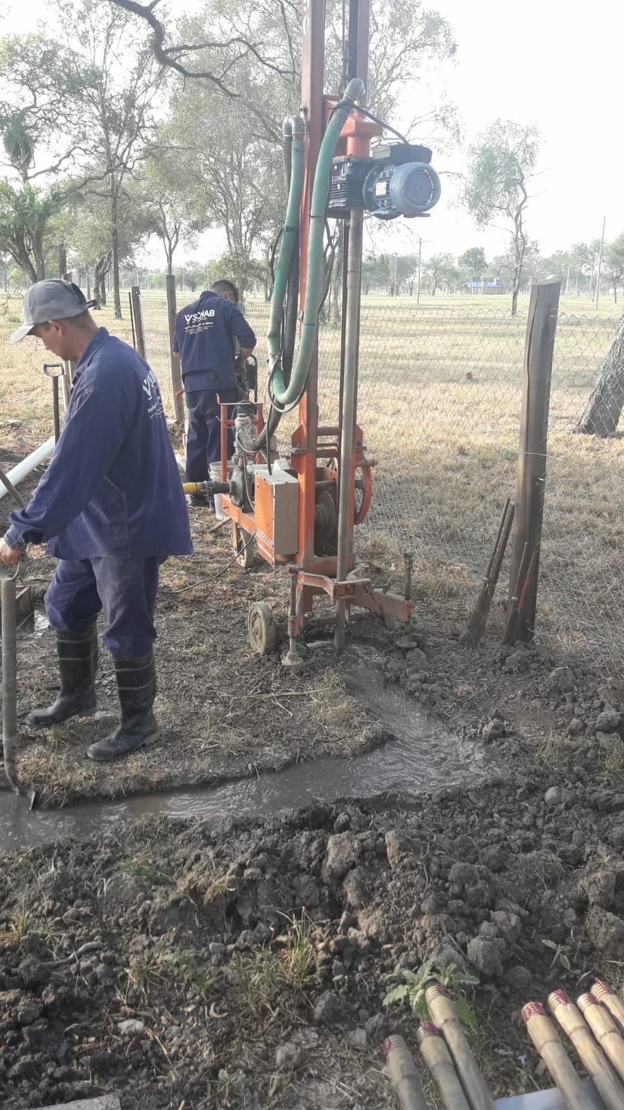 Trabajadores perforando un pozo junto a un campo fangoso, con una plataforma portátil y tuberías apiladas.
