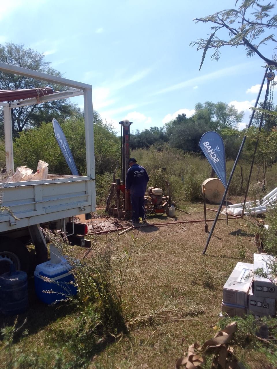Lugar de trabajo al aire libre con un camión, dos personas, tiendas de campaña y suministros apilados en un campo de hierba.
