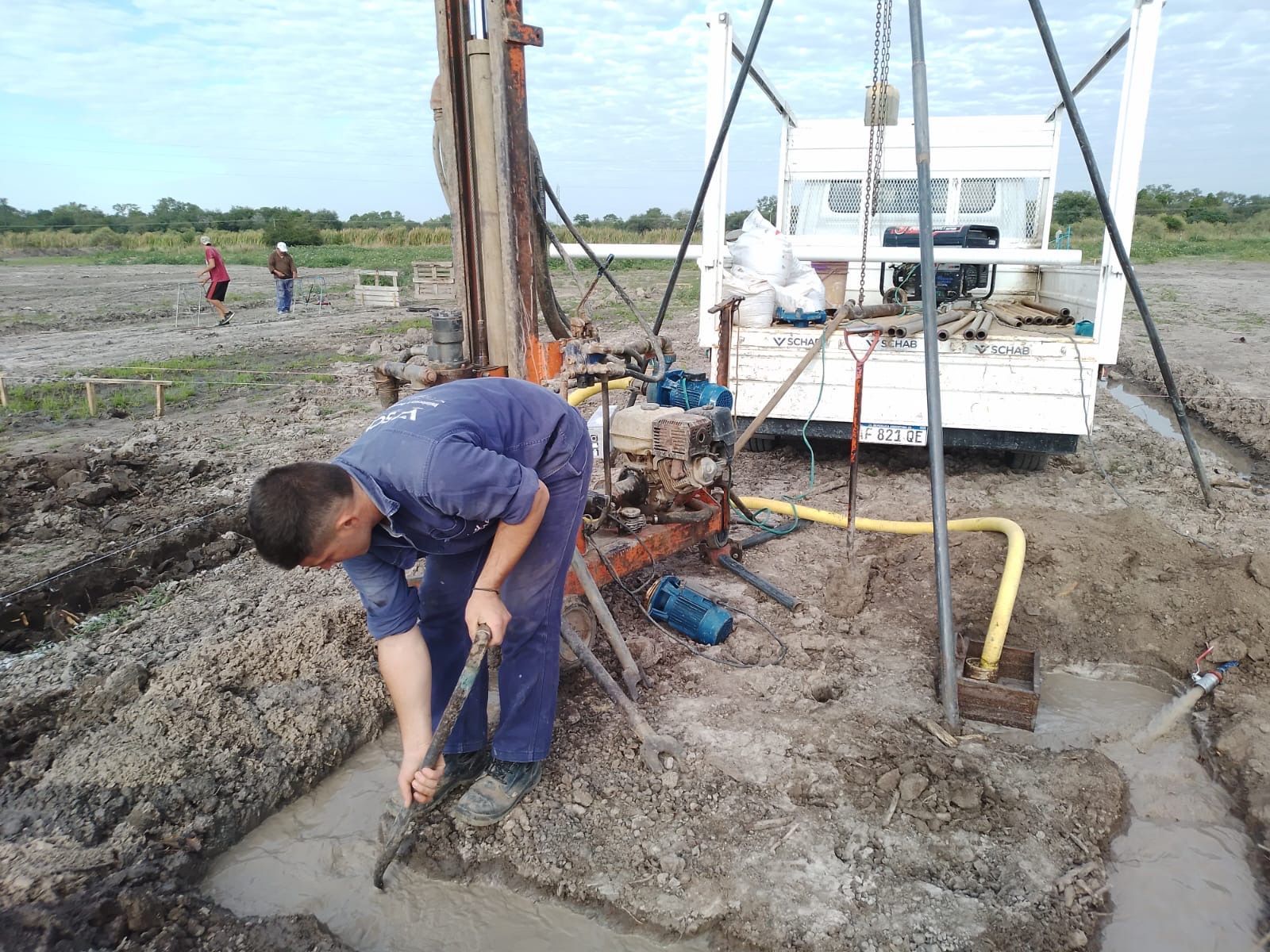 Un trabajador perfora un terreno fangoso junto a un camión blanco y una plataforma de perforación en un campo húmedo.