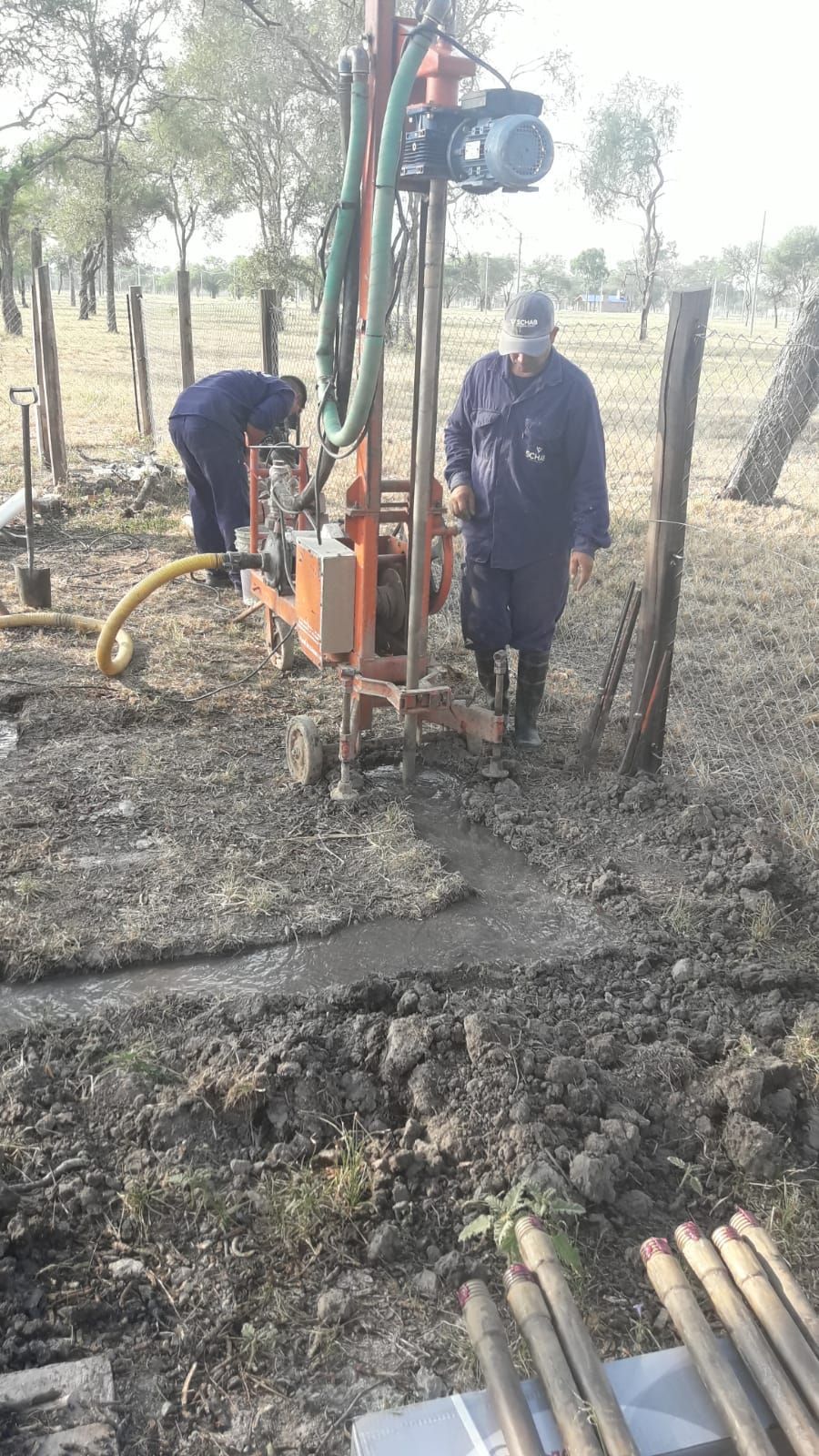 Dos trabajadores manejando una plataforma de perforación junto a postes de una cerca en un campo rural fangoso.