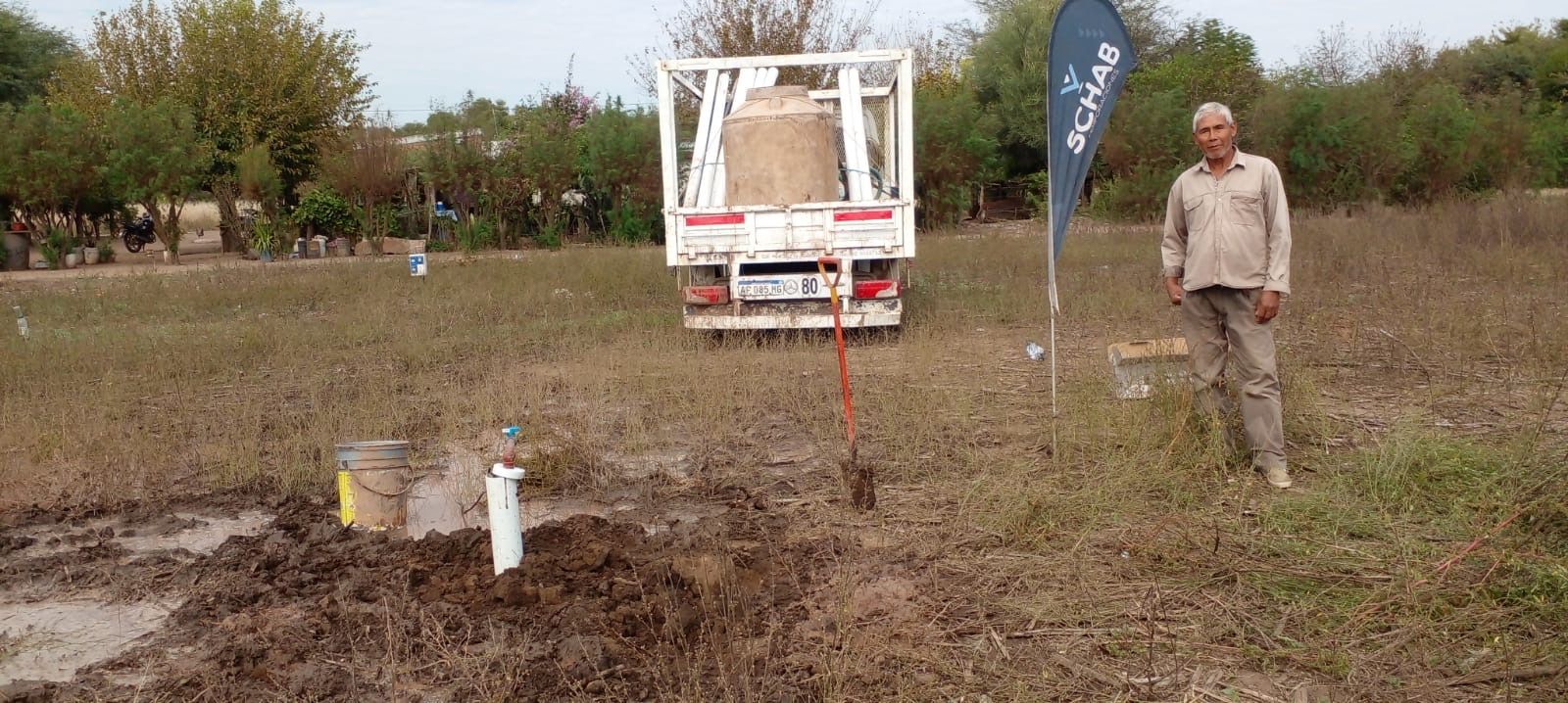 Camioneta blanca en un sitio de construcción embarrado con banderas, tuberías y un hombre de pie cerca.