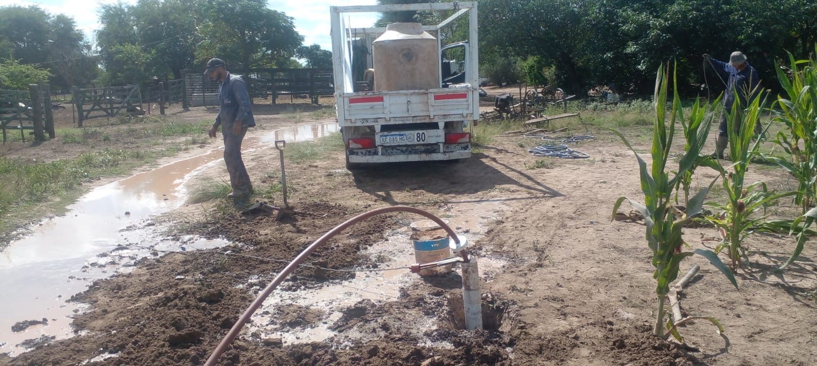 Un trabajador junto a un camión blanco rociando agua sobre un terreno fangoso en un campo rural.