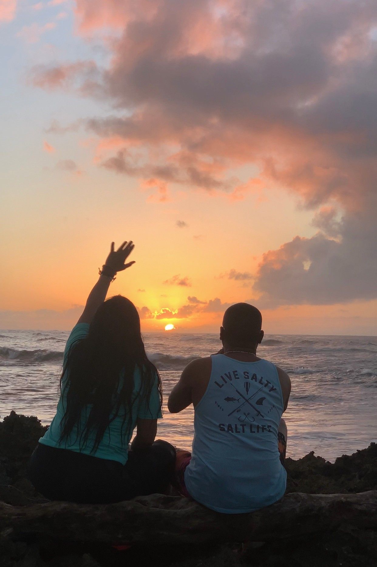 A man and a woman are sitting on a rock at the beach watching the sunset.