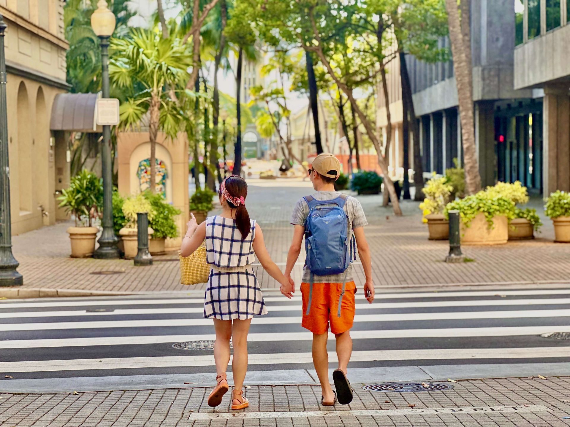 Two children holding hands walk across a crosswalk, buildings and greenery in background.