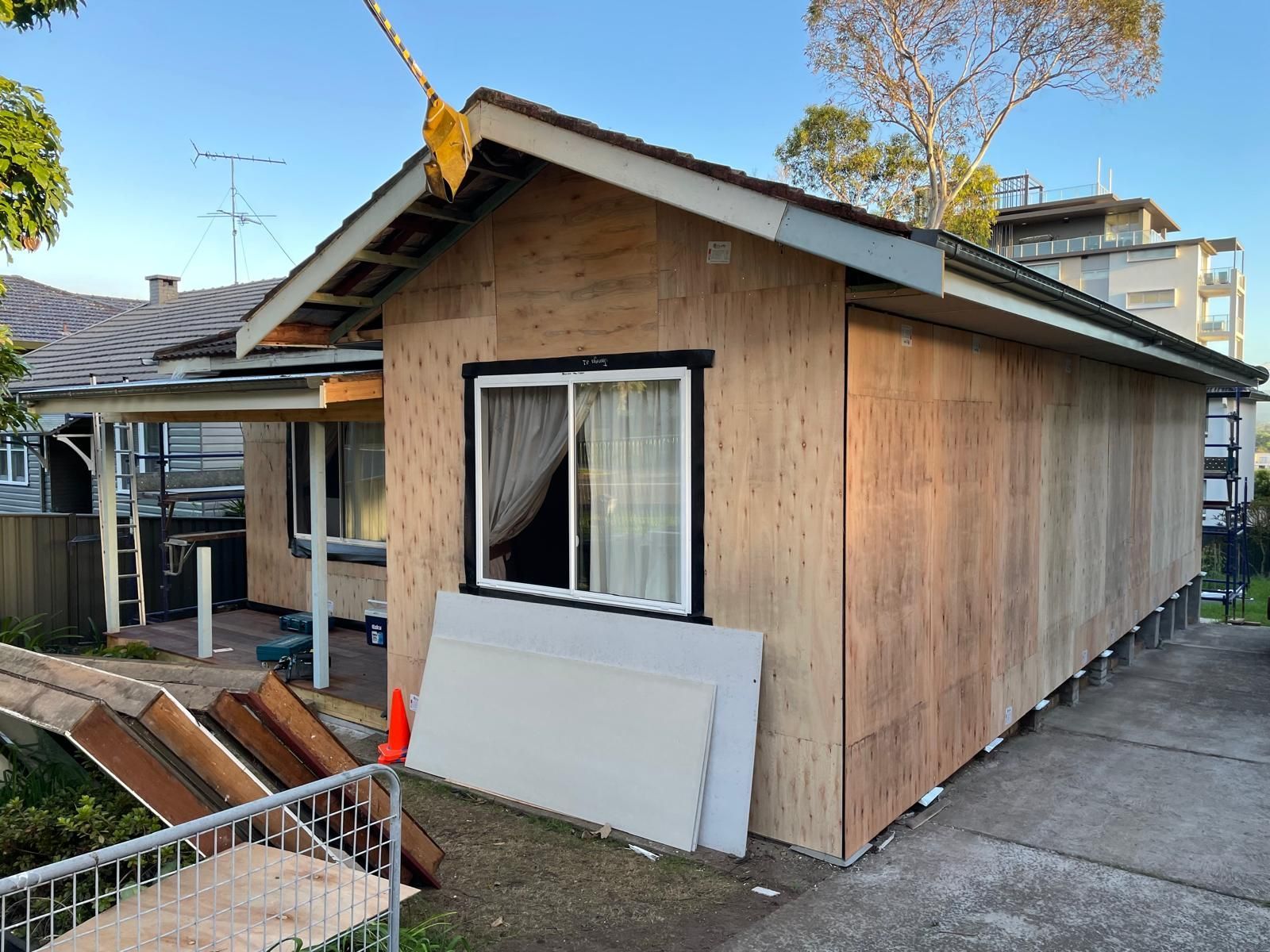 House exterior under construction, plywood siding, window with curtain, sunny day.