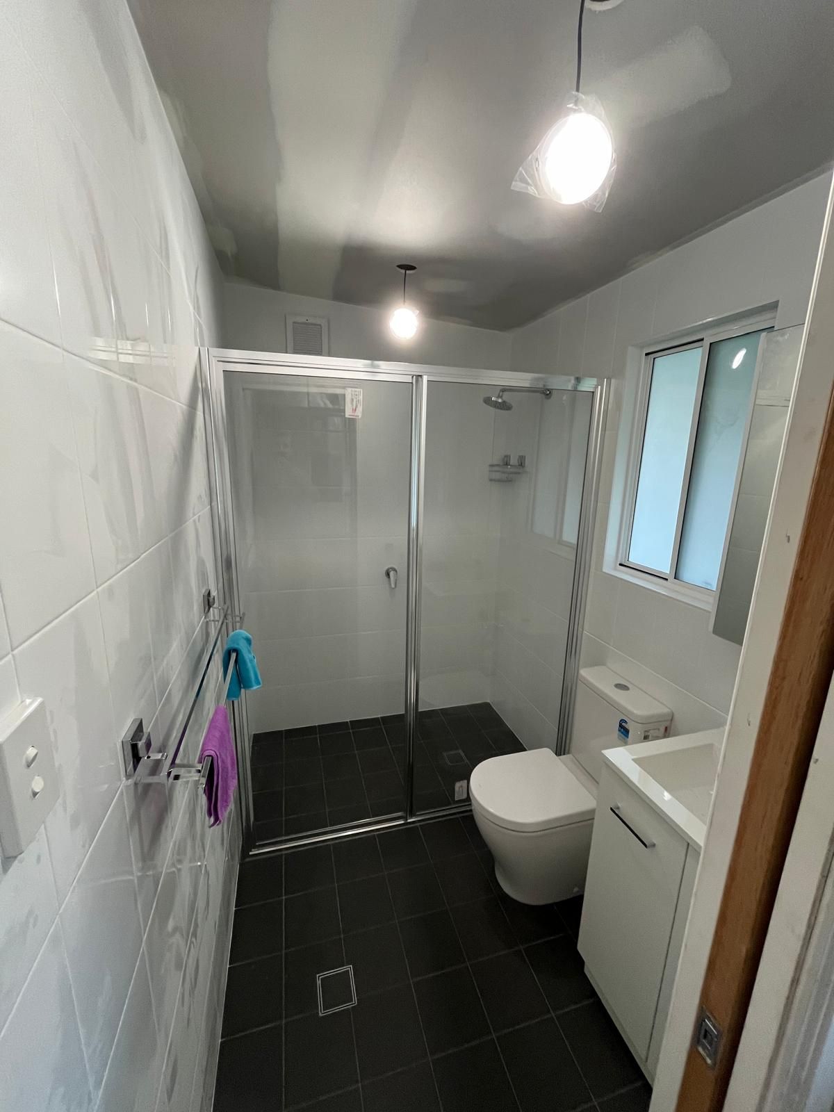 A small, modern bathroom with white tiles, a glass shower, a toilet, and a sink; dark floor.