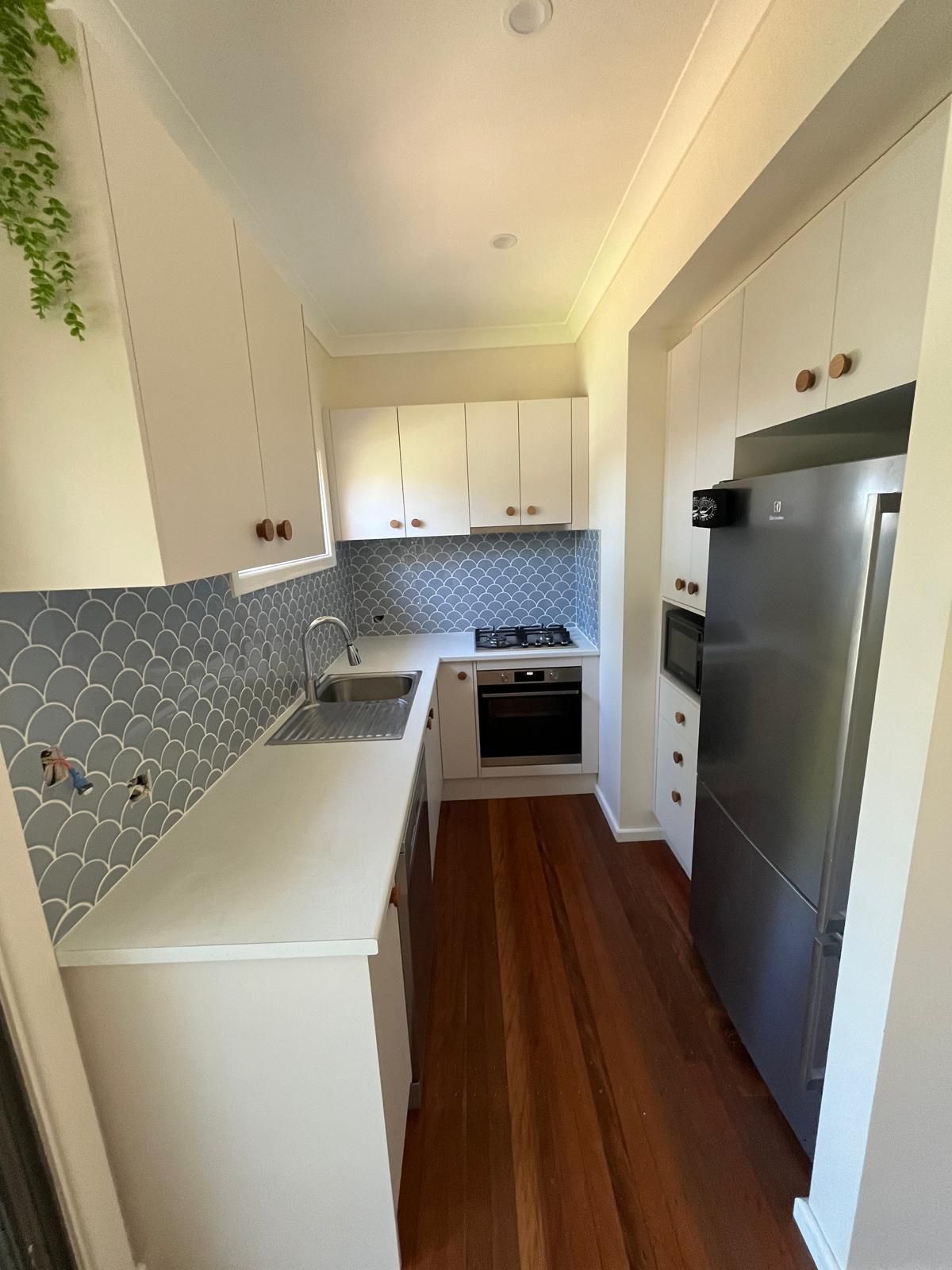 Narrow kitchen with white cabinets, wood floor, blue tile backsplash, stainless steel fridge.