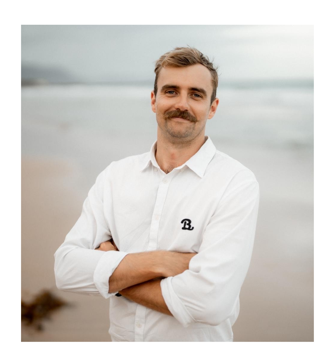 Man with mustache, arms crossed, stands on a beach, wearing a white button-down shirt.