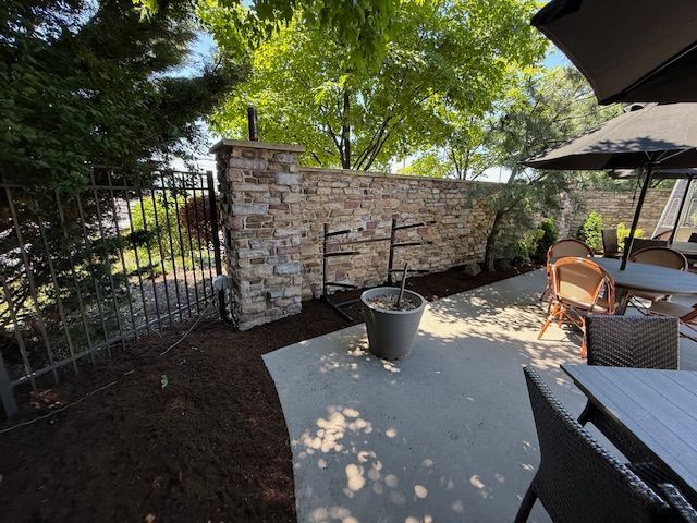 Shaded patio with stone wall, metal gate, potted plant, and outdoor table and chairs under trees