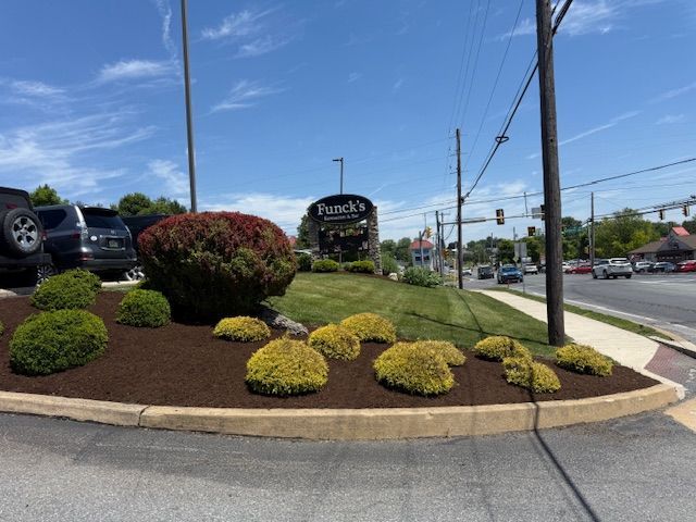 Landscaped roadside entrance with trimmed shrubs, flowers, and a sign by a busy street under a blue sky