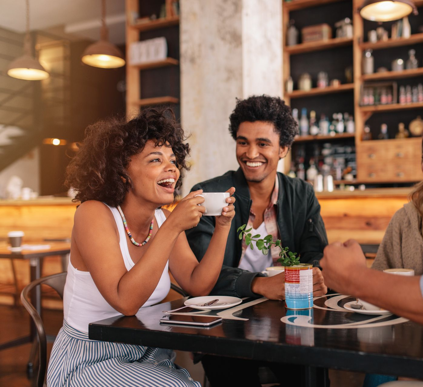 Two people laughing at a table in a cafe, one holding a cup.