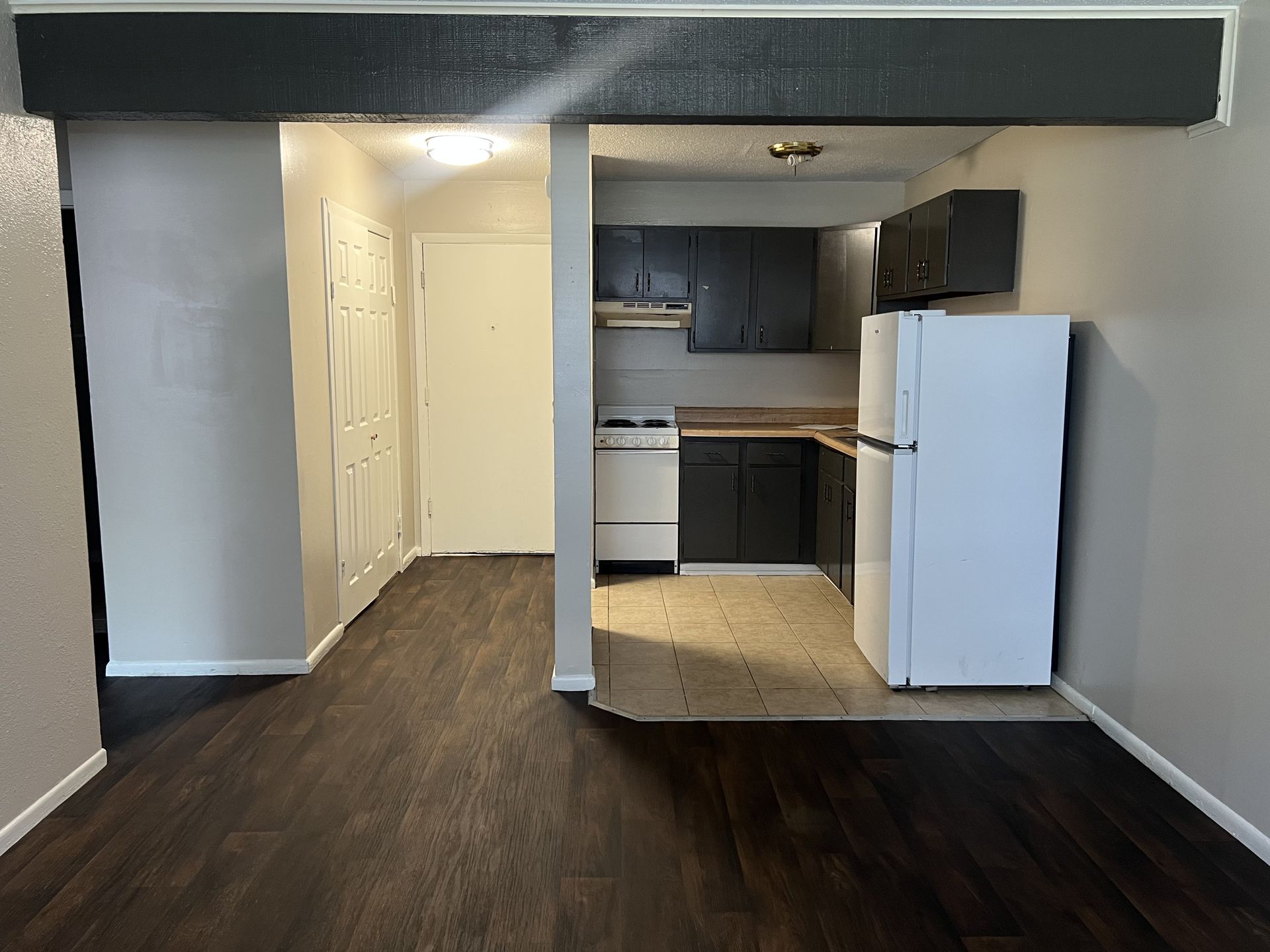 Interior view of a kitchen and adjacent room, with dark wood flooring, and black cabinets.