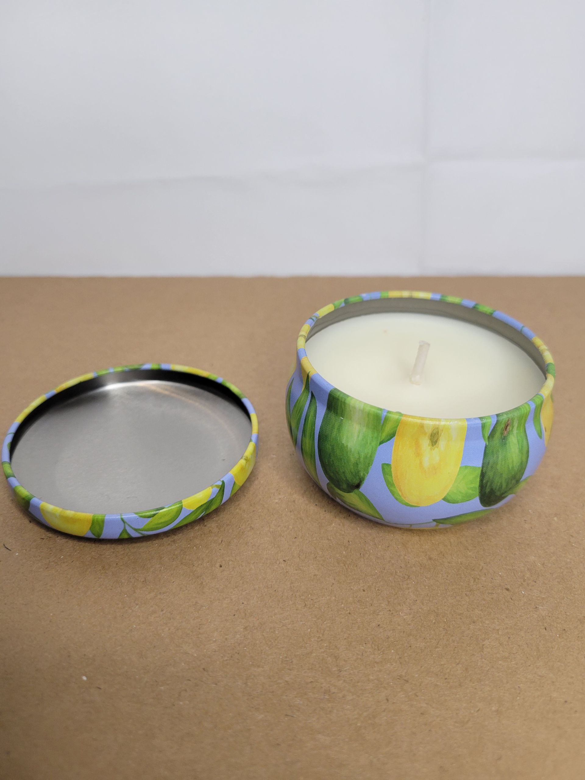 Two floral candle jars, one lid off beside a lit white candle on a brown surface.