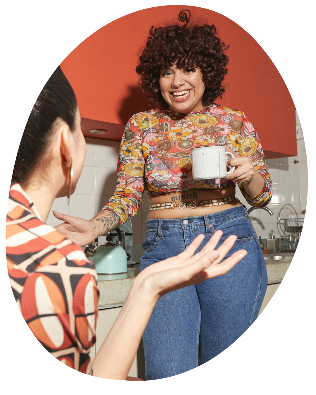 Person in a floral top offers a mug in a kitchen, smiling at someone in the foreground.