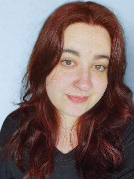 Portrait of a person with wavy red hair and a slight smile against a light blue background
