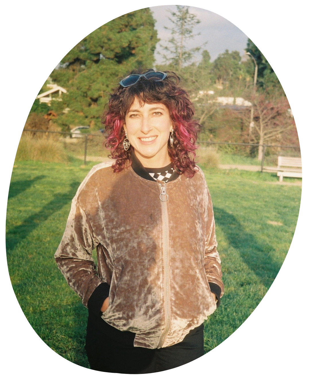 Person standing outdoors in a brown sweater and black pants, smiling in a grassy park.