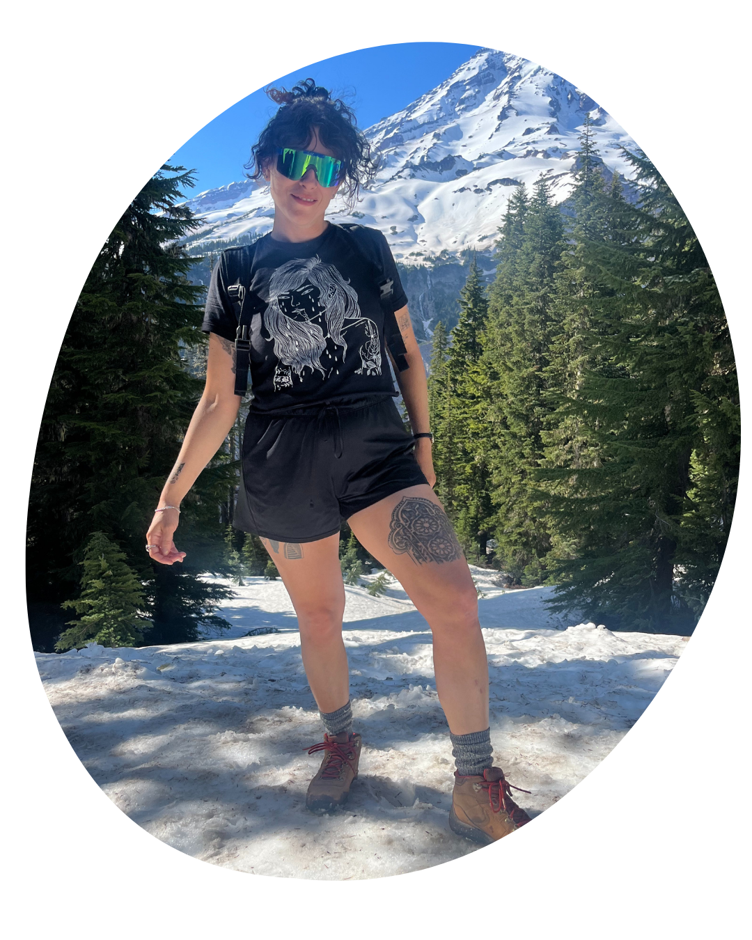 Person standing on snowy mountain trail with blue hair, sunglasses, and shorts, trees and a snow-covered peak behind.