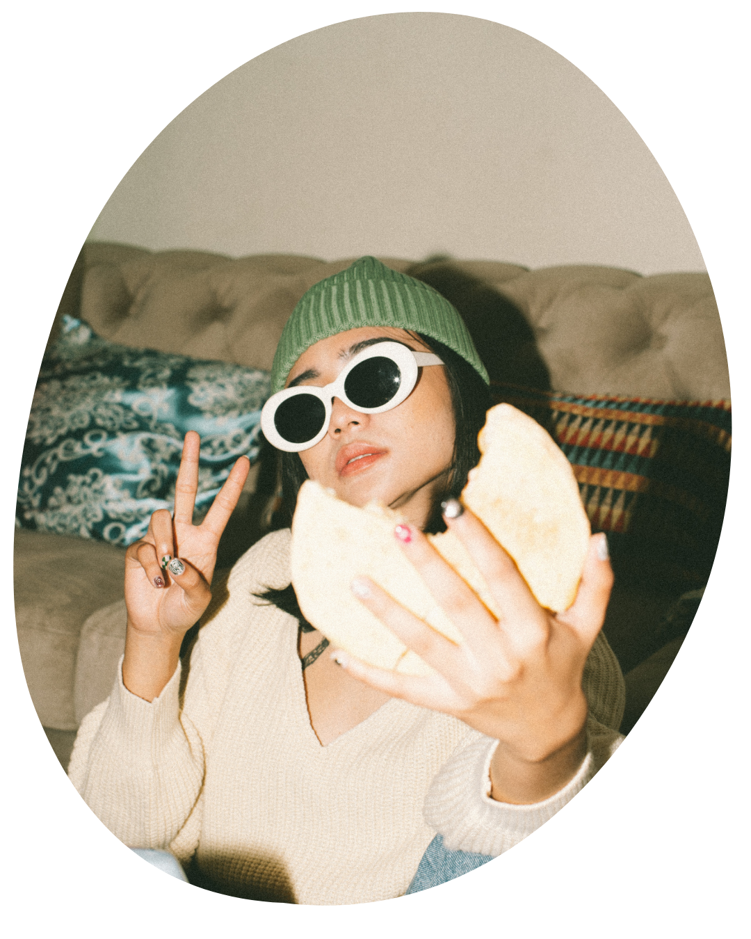 Person in green beanie and white sunglasses taking a selfie, making a peace sign on a couch.