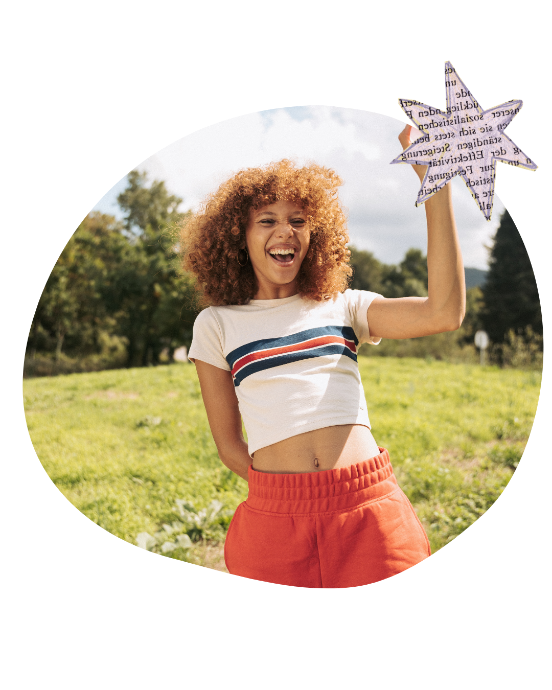 Smiling woman in a striped crop top and orange pants standing in a grassy field, holding a star-shaped sign.
