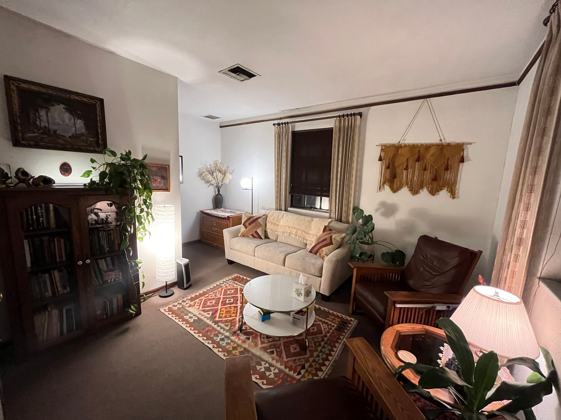 Cozy living room with beige sofa, patterned rug, dark cabinet, plants, and warm lamp light
