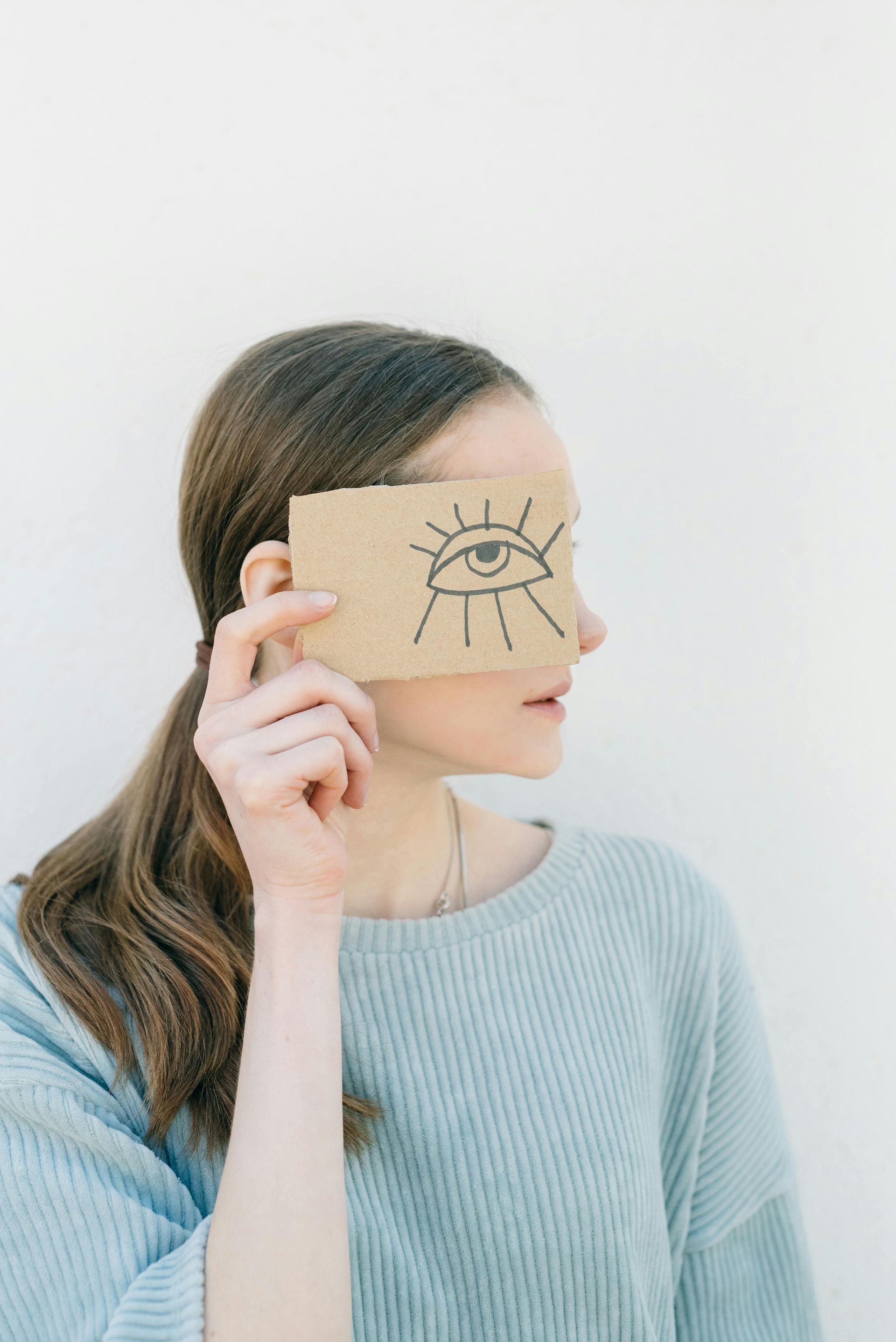 Person holding a cardboard card with a simple eye drawing over one eye against a plain light background