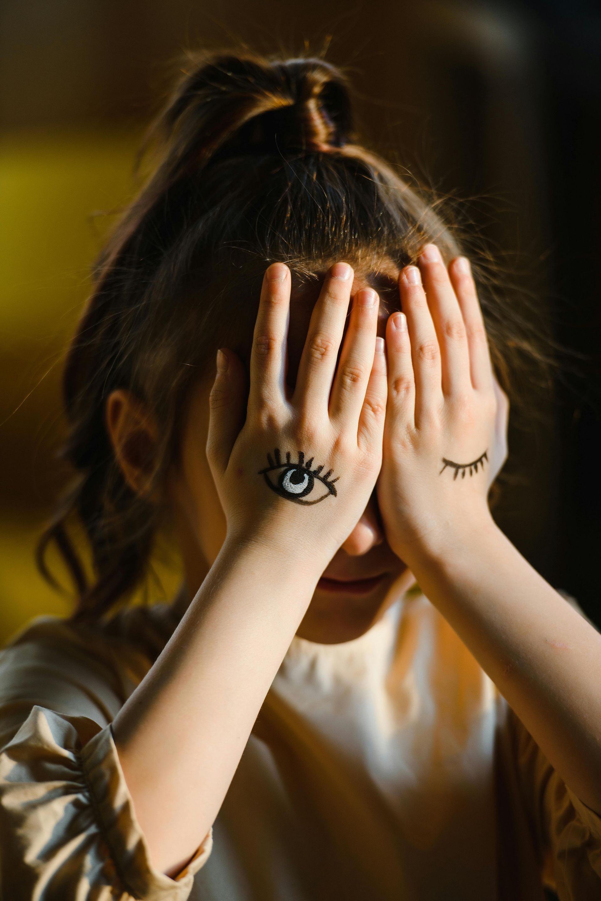 Child covering face with hands, drawn eyes on palms, in warm indoor light.
