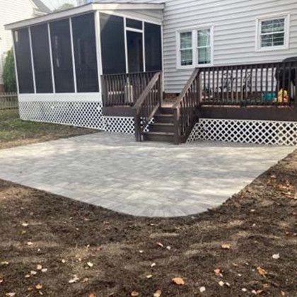 A gray paver patio adjoins a deck and screened porch. White lattice and brown stairs are visible.