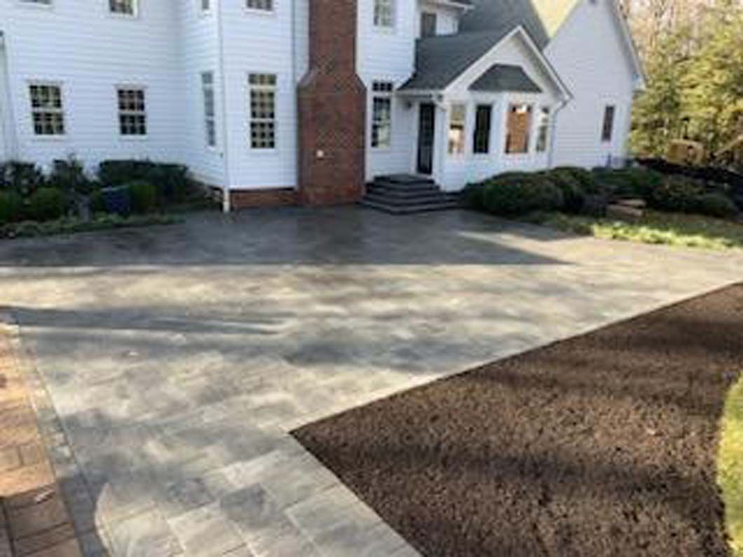 Paved driveway leading to a white house with a brick chimney. Soil bed and lawn on the right.