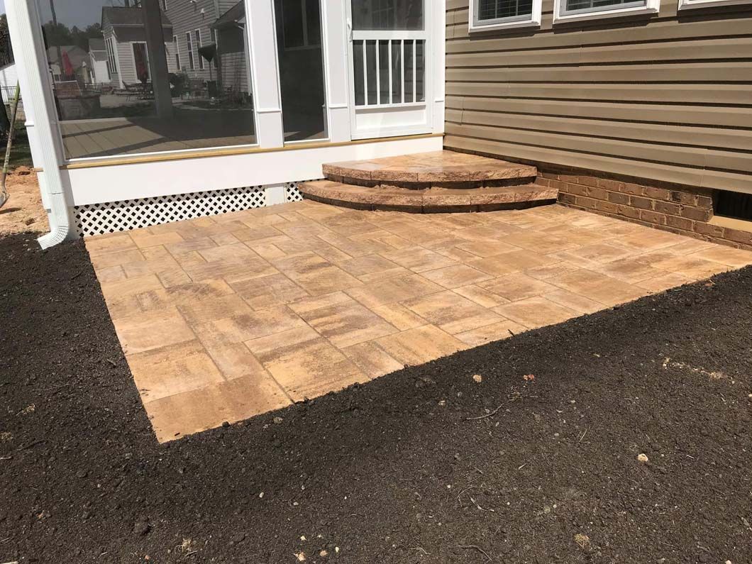 Brown paver patio with two steps leading to a porch entrance, next to a tan siding house.