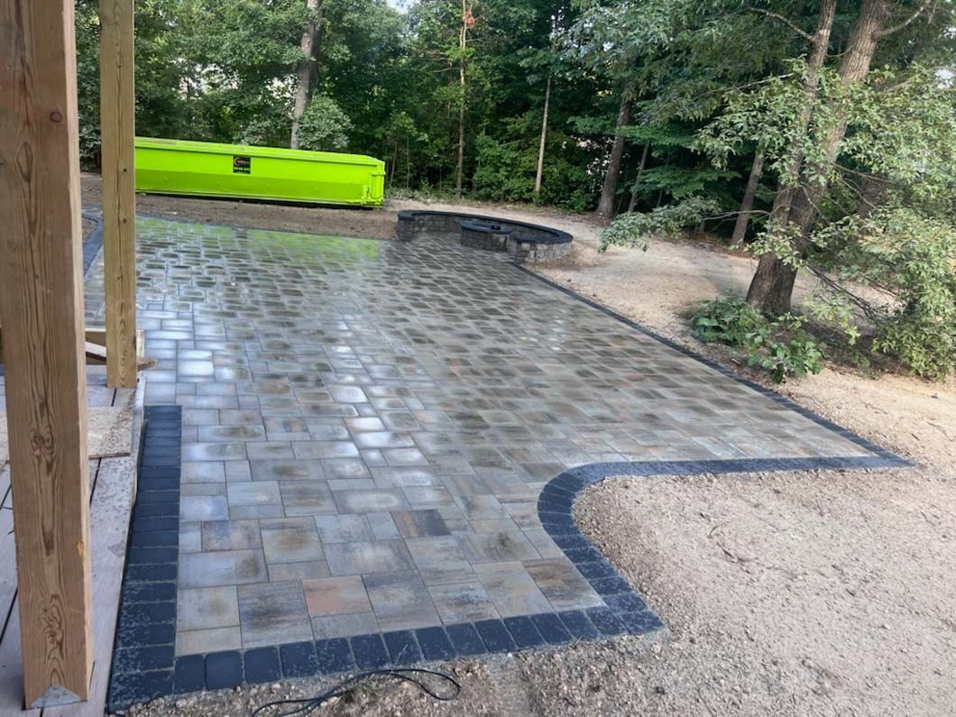 Patio paved with gray and brown stones, bordered with dark stones, next to a wooded area.
