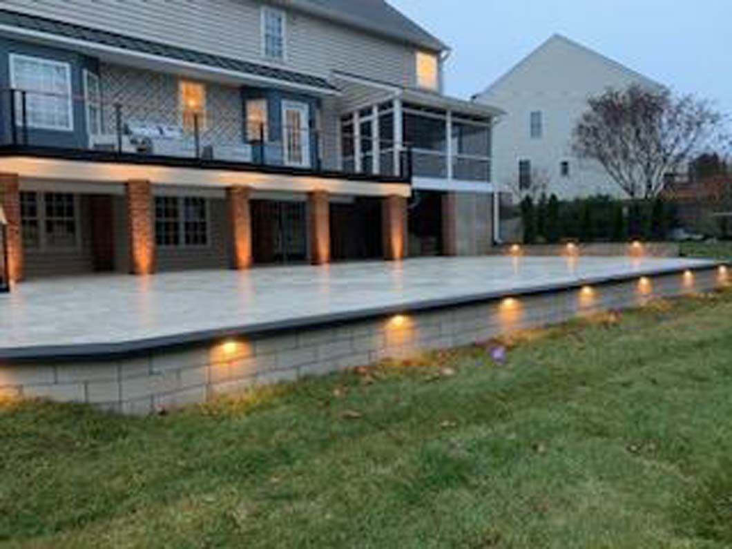 Backyard patio with retaining wall, lit by ground-level lights. The house has a deck, screened porch, and brick columns.