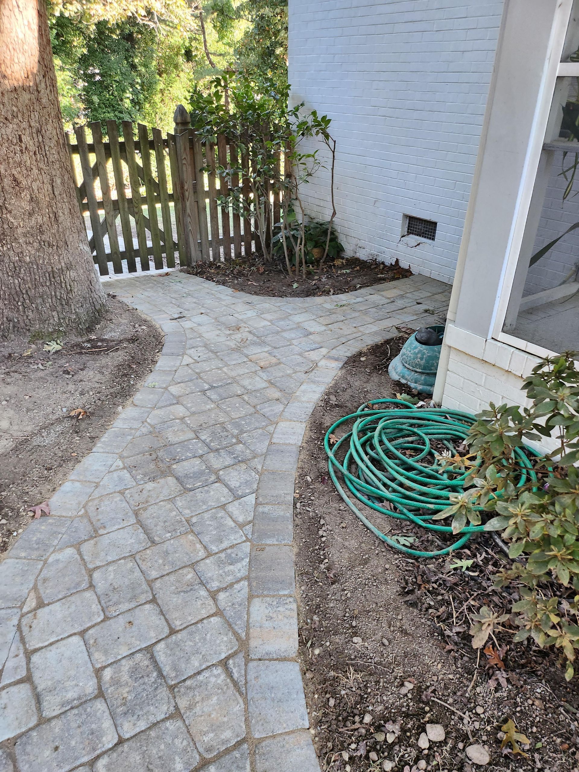 Brick pathway curves past a tree and green garden hose, leading to a wooden gate.