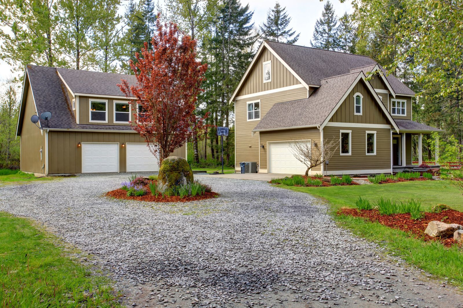 Two-story house with detached garage, gravel driveway, and red tree, set in a wooded area.
