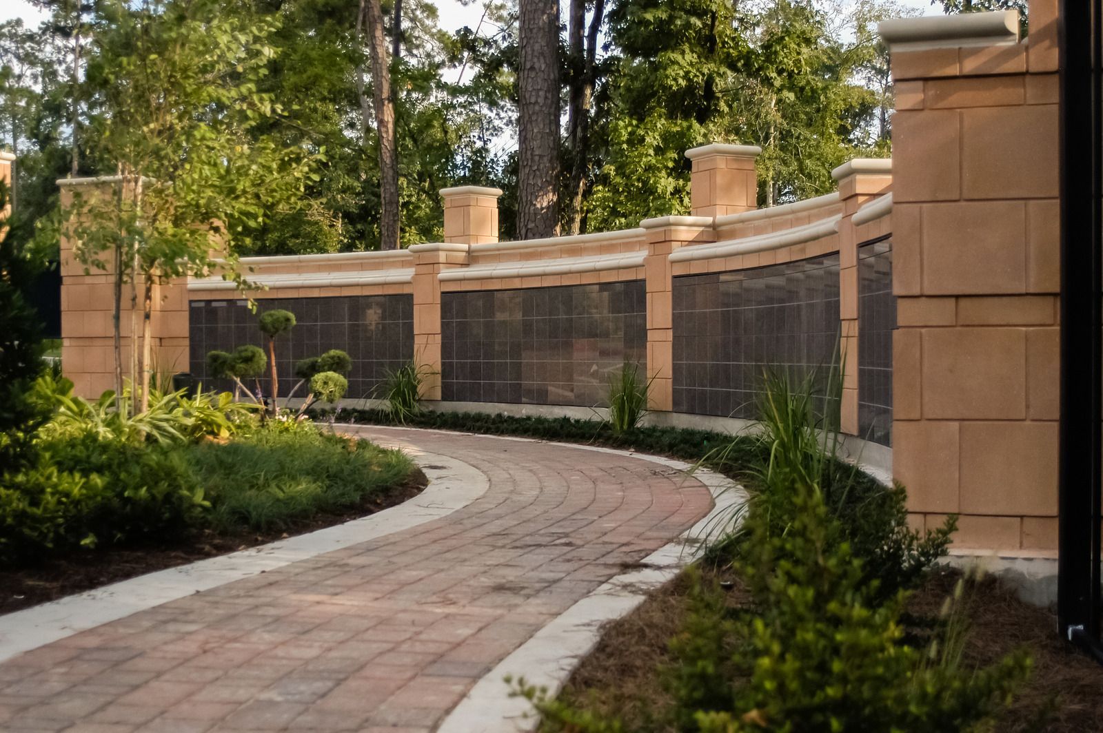 Brick pathway curves past a wall of bronze plaques, bordered by landscaping and trees.