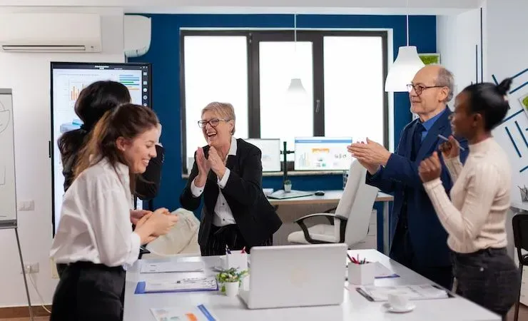 Team meeting in a bright office, with colleagues around a table discussing and clapping near laptops and papers.