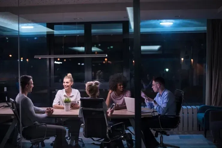 People meeting around a table in a dimly lit office at night, with laptops and a plant centerpiece.