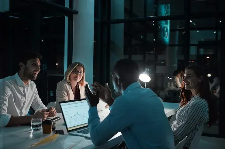 Team meeting in a glass-walled office, with coworkers around a laptop under soft evening light