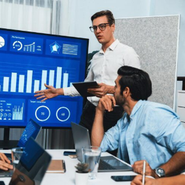 Team meeting in office analyzing blue dashboard charts on a large monitor