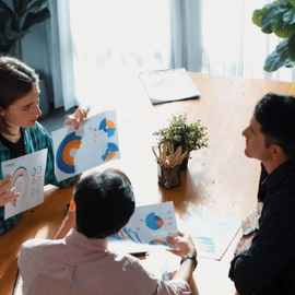 Three people discuss colorful charts at a table in a bright office